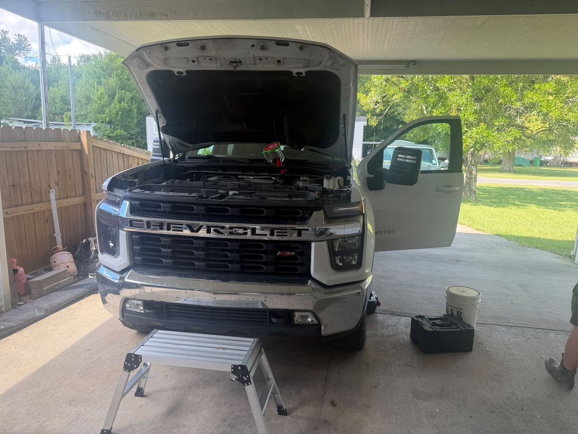 A full-size Chevrolet pickup with the hood up in a carport, with a step stool and drain pan set out for a mobile oil change