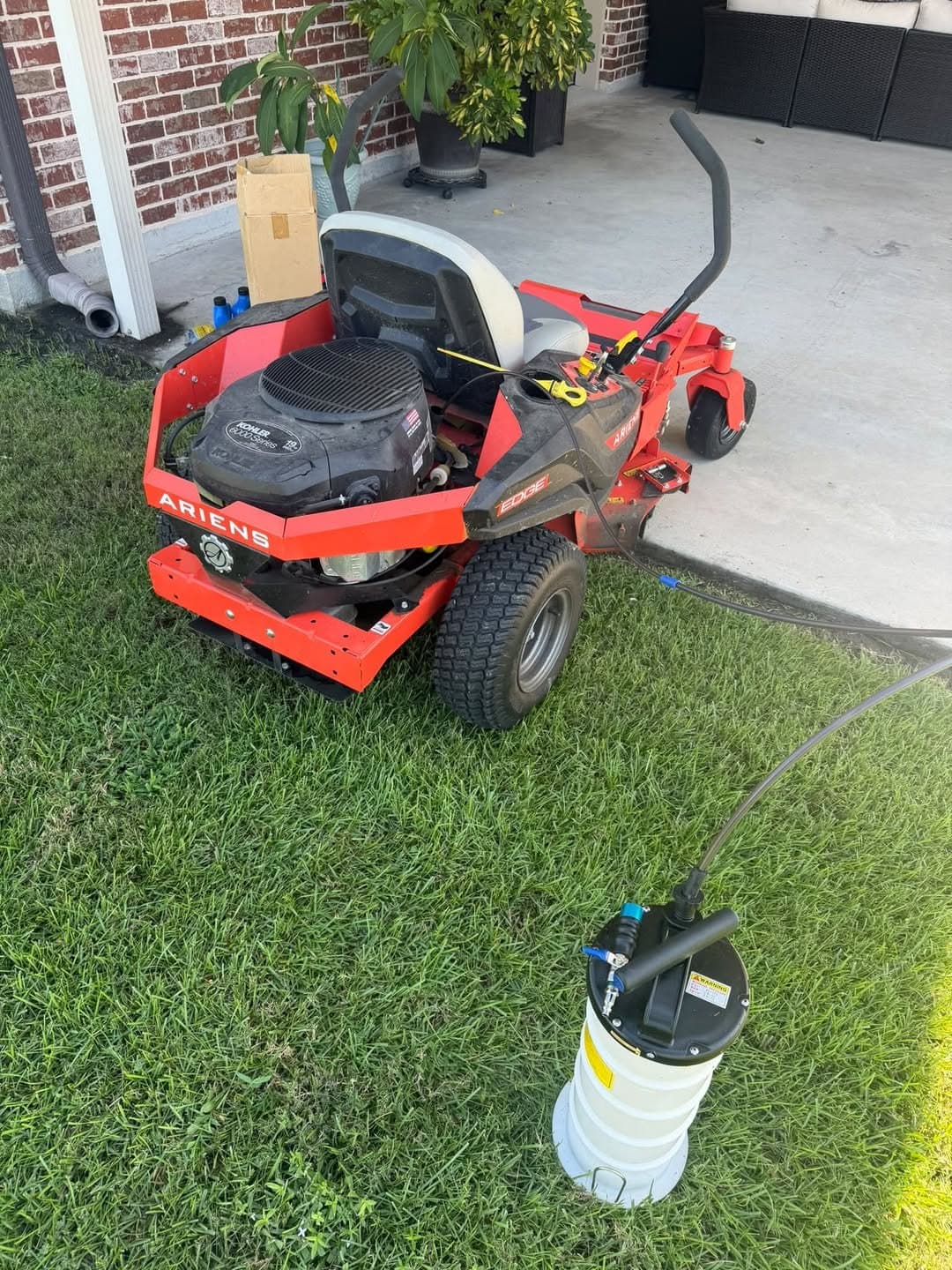 A red Ariens zero-turn mower on the grass with a manual oil extractor connected, drawing the old oil out