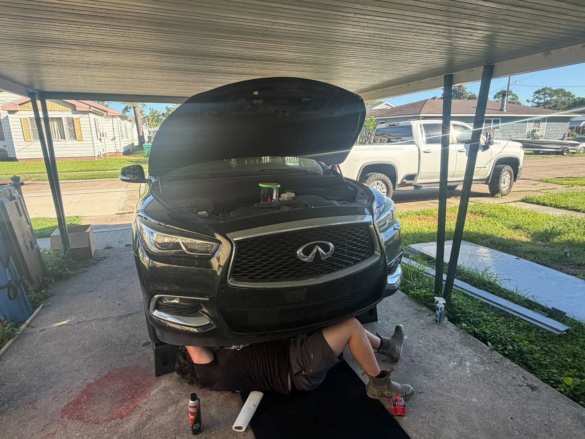 A dark SUV with the hood up under a carport while a Bayou Boyz technician works underneath it, with a drain pan and oil set out on the ground