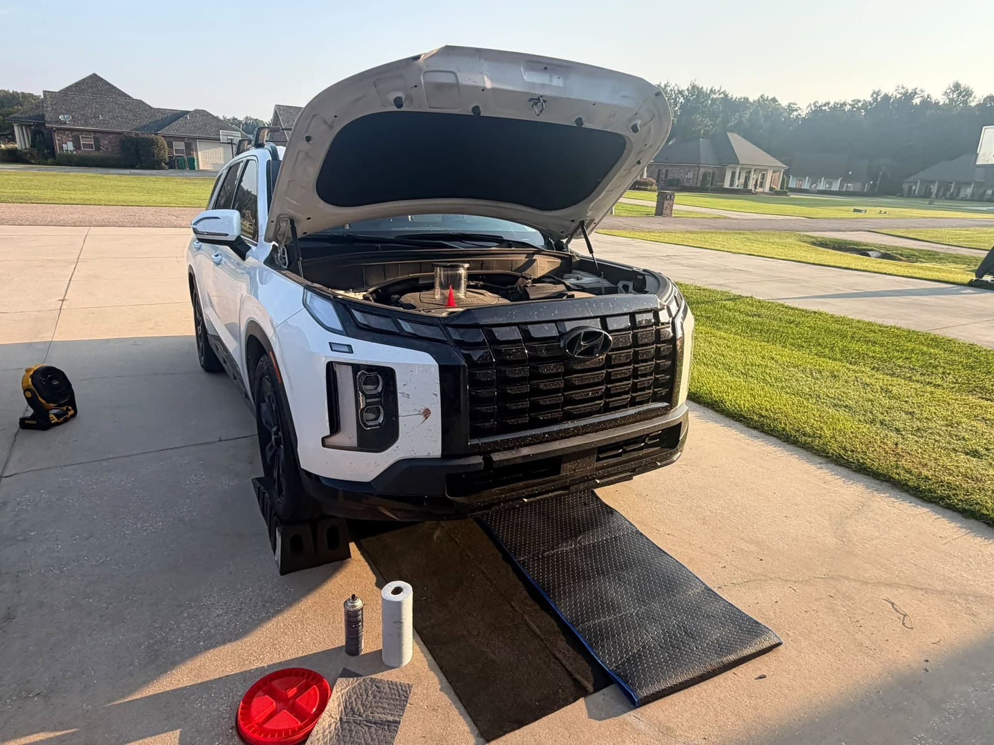 An SUV up on ramps in a Louisiana driveway with the hood open and a drain pan, oil and paper towels set out for a Bayou Boyz mobile oil change